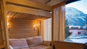 A cozy wooden cabin room with a sofa and a table, a window with curtains, and a view of snow-covered mountains. - 50m from ski slopes, Ground floor, view mountain, parking, tv, ski locker, 18m², Montgenèvre (Montgenèvre)