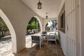Un patio cubierto con suelo de piedra, paredes blancas y arcos. Hay dos sillas y un banco.