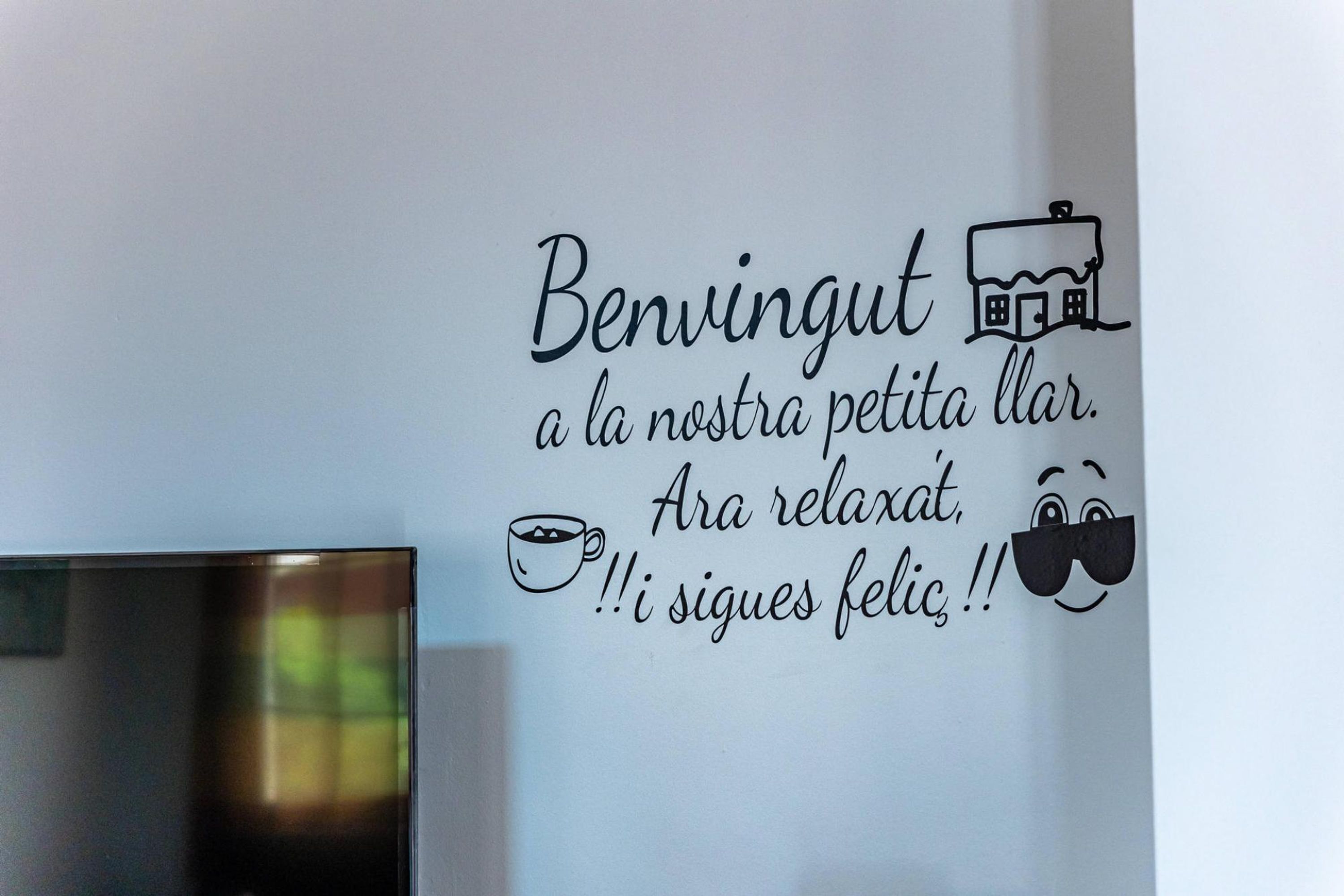 Paret amb un senyal decoratiu que representa una tassa de cafè i ulleres de sol, i text en una llengua estrangera.