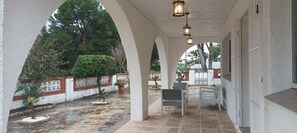 Un patio cubierto con suelo de piedra, paredes blancas y arcos. Hay dos sillas y una mesa, con plantas en macetas y una pequeña fuente.