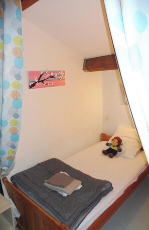 Ein Einzelbett mit Holz-Kopfteil, ein kleiner Nachttisch, ein an der Wand hängendes Bild und ein Vorhang mit Punkten.
