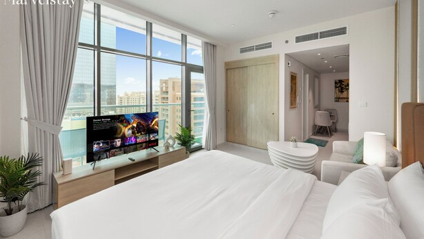 Ein modernes Hotelzimmer mit einem großen Bett, einem Fernseher auf einer Holzkonsole, Blick auf die Stadt und einer separaten Sitzecke mit einer Lampe.