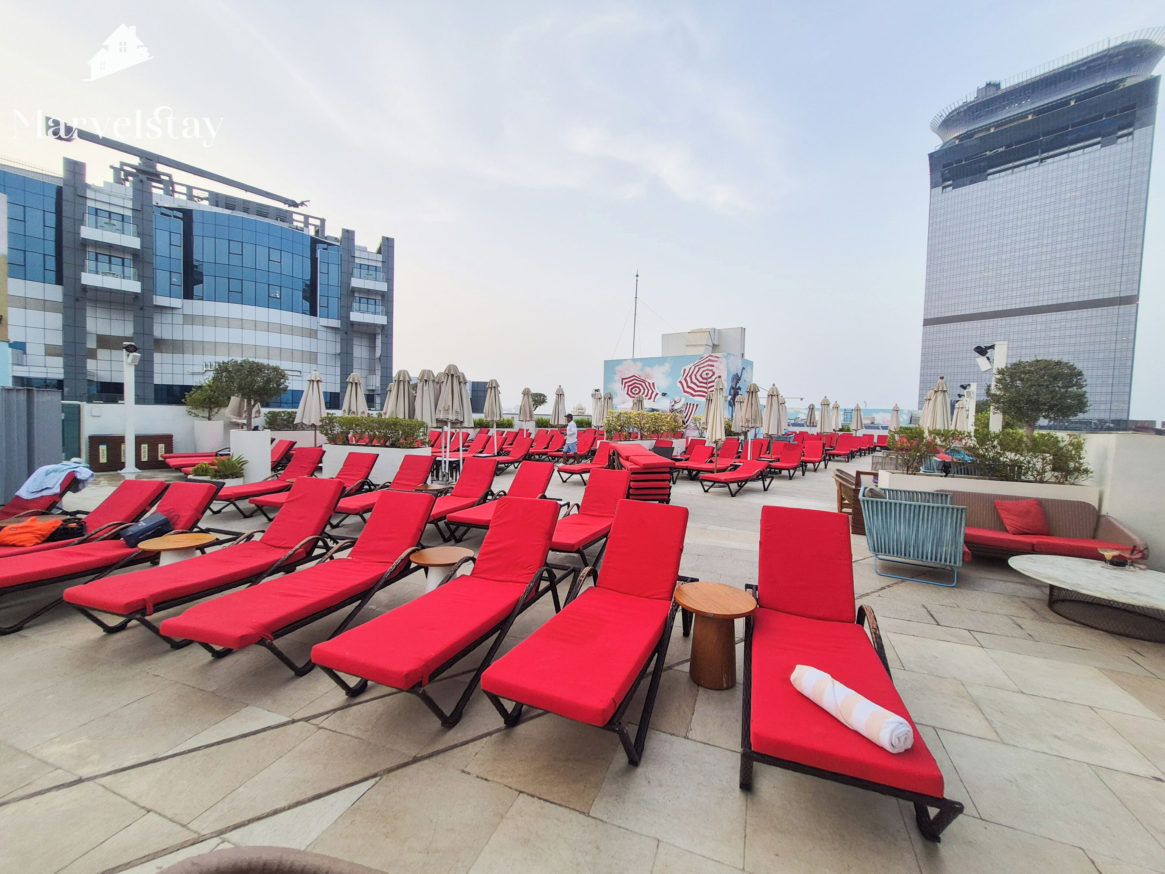 Un espace de détente sur le toit, avec des fauteuils rouges et des parasols, une vue sur des bâtiments modernes et un ciel dégagé.