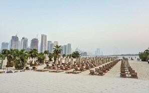 Une plage bordée de rangées de chaises longues et de parasols, avec des palmiers, et un paysage urbain en arrière-plan, où se dressent de hauts immeubles.