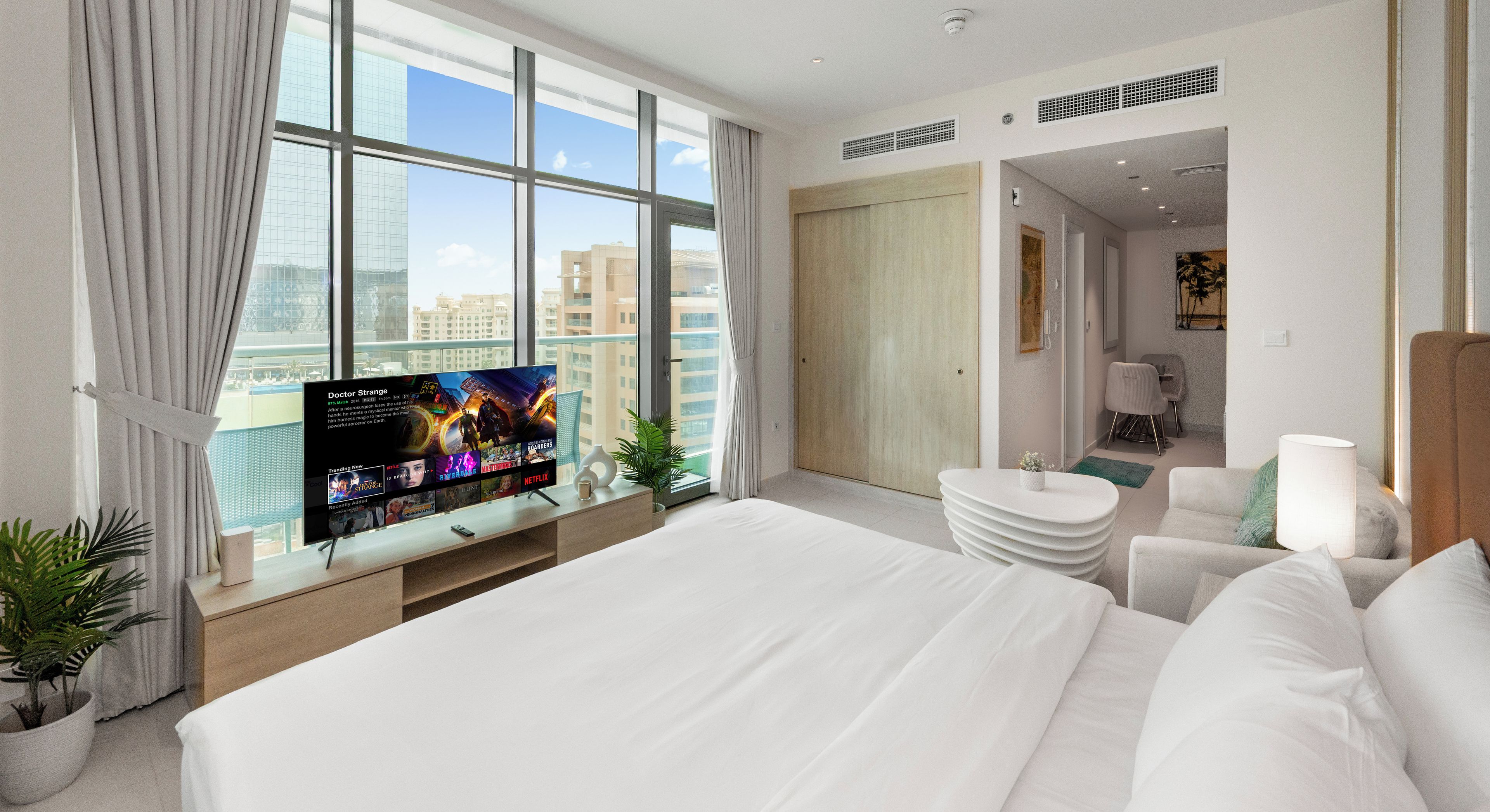 Una habitación de hotel moderna con una cama grande, un televisor sobre una consola de madera, vistas a la ciudad y una zona de estar separada con una lámpara.