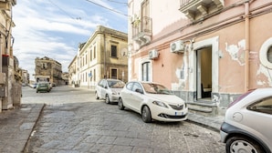 Calle empedrada flanqueada por edificios históricos, coches estacionados y una entrada visible a un edificio con un letrero.