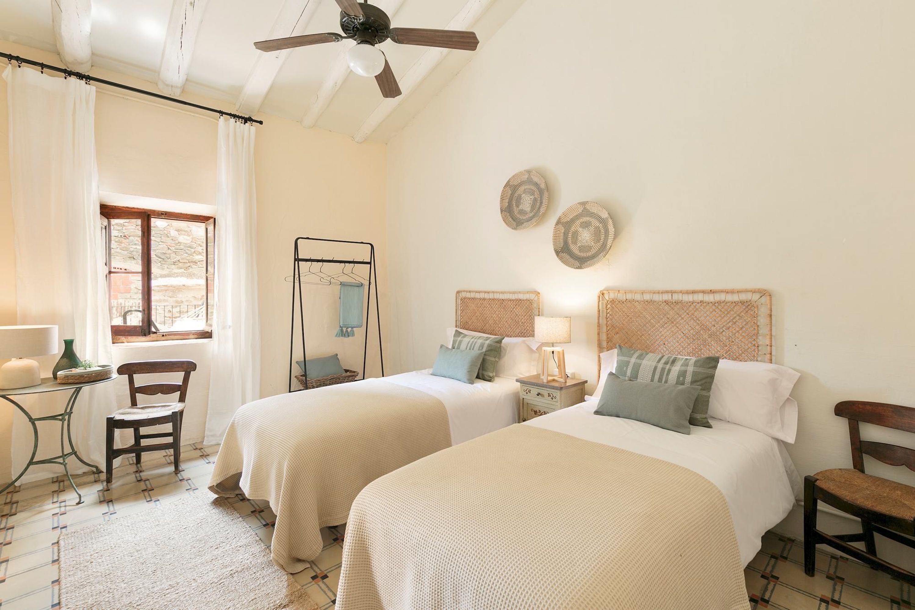 Une chambre avec deux lits, un ventilateur de plafond, une fenêtre avec des rideaux, une petite table avec une lampe et une chaise.
