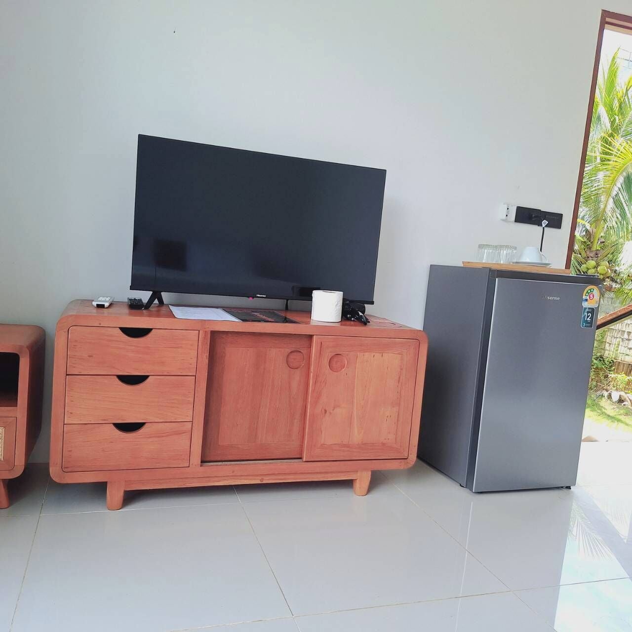 A room with a flat-screen TV on a wooden cabinet, a small refrigerator, and a window with a view of greenery.