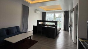 A modern living room with a sofa, coffee table, and a desk with a chair. There is a large window with curtains, a ceiling with recessed lighting, and a door on the right. - The wide view resort cafe (Thep Krasattri)