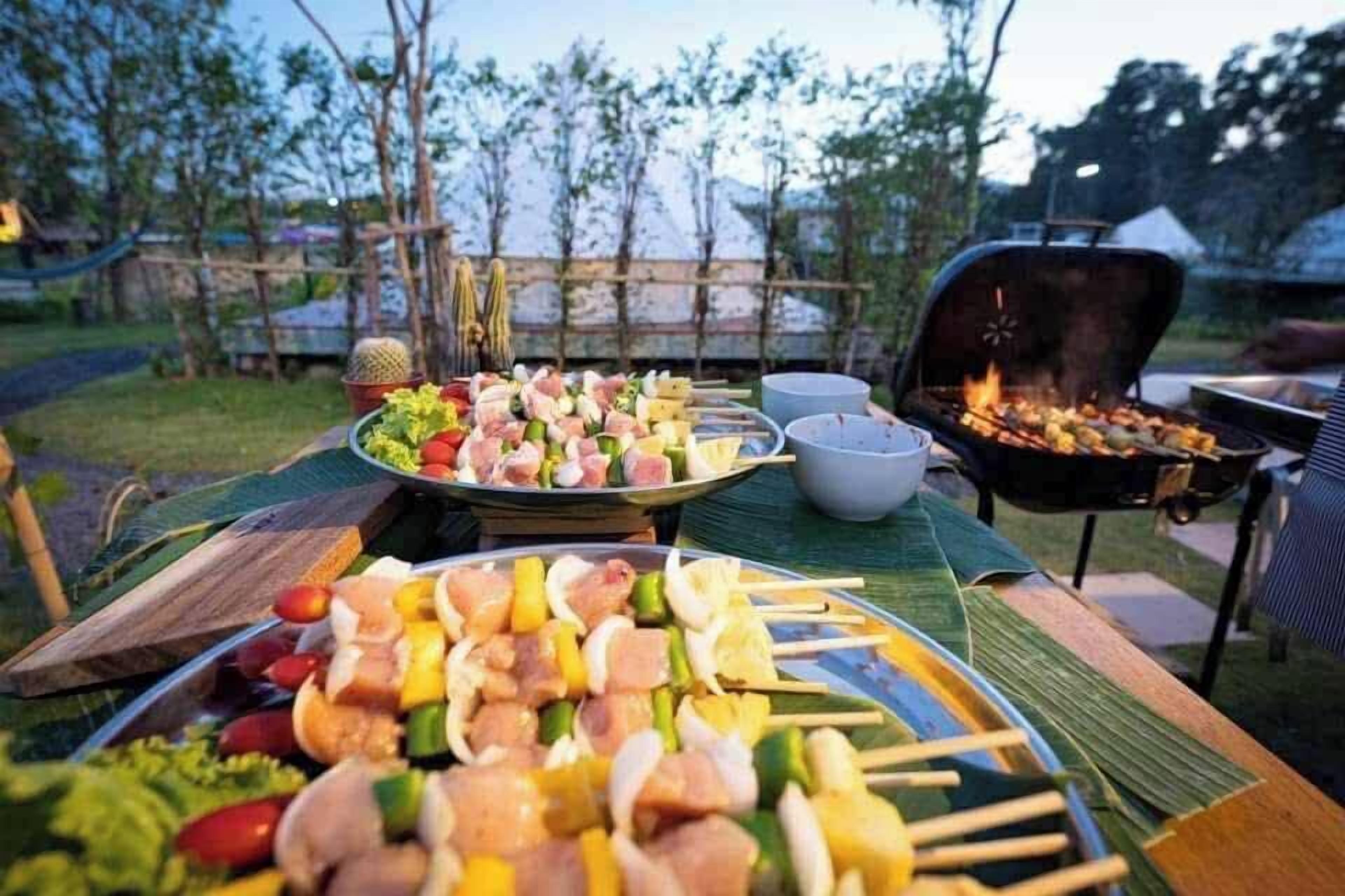 Un coin barbecue en plein air avec des brochettes de viande et de légumes, un gril et une table à manger recouverte d’une nappe verte.