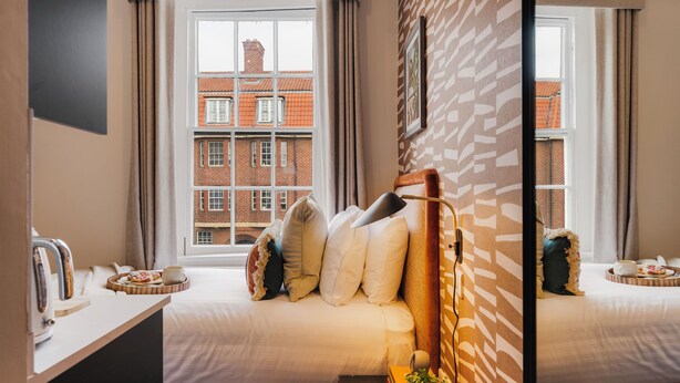 Une chambre d’hôtel avec deux lits, une petite table avec des éléments pour le petit-déjeuner, une plante et une vue sur des bâtiments en briques rouges, visible à travers de grandes fenêtres.