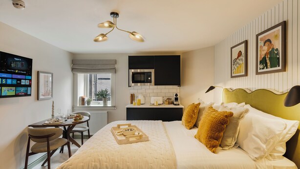 A hotel room with a bed, a dining table with chairs, a kitchenette, and a TV mounted on the wall.