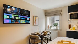 A modern hotel room with a flat-screen TV displaying various channels, a dining table with a meal, a bed with a tray, and a window with a view.