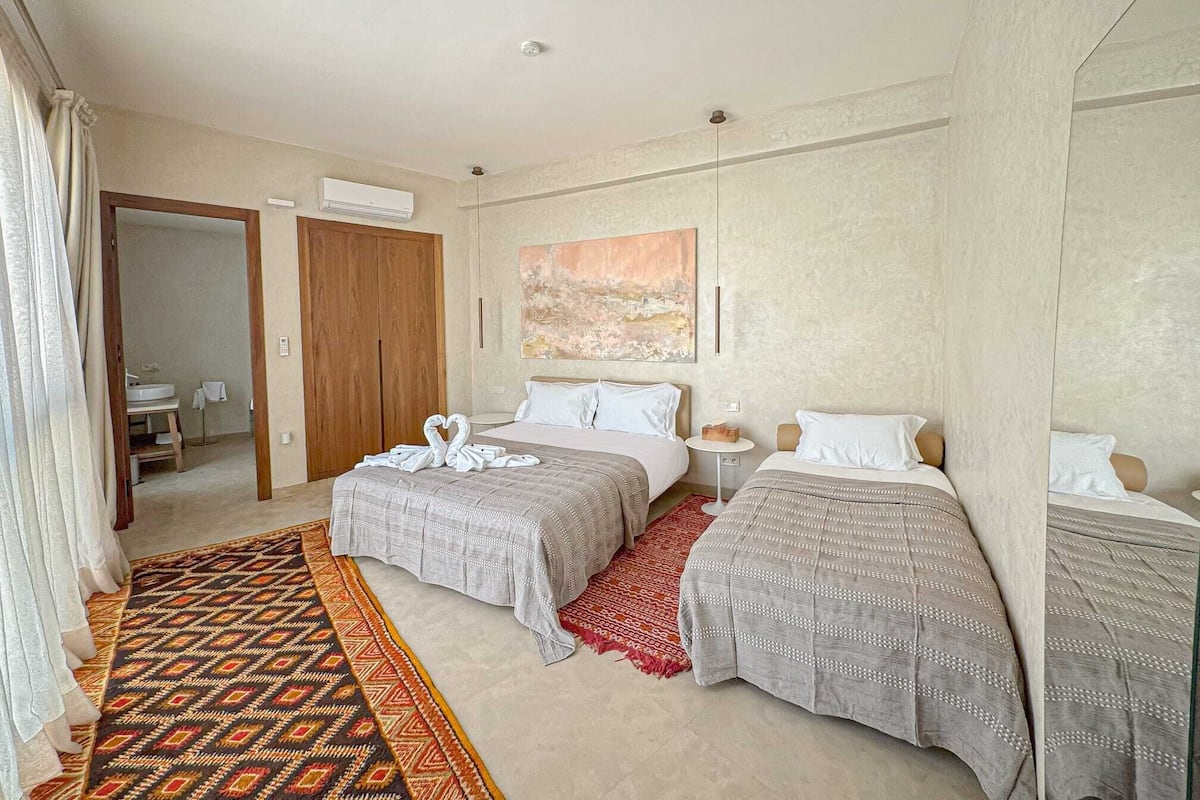 A hotel room with two beds, a large window, a wooden wardrobe, and a red patterned rug.