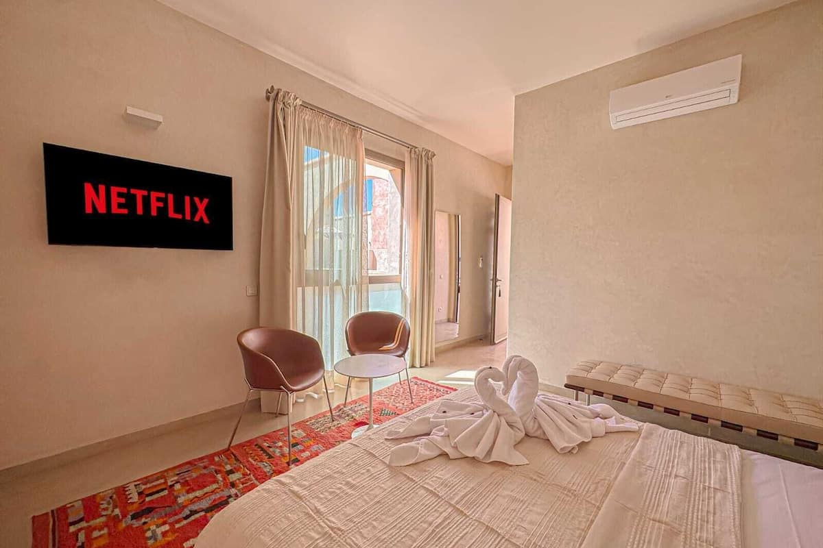 A hotel room with a bed, a TV mounted on the wall, a window with curtains, a small table, and a chair.
