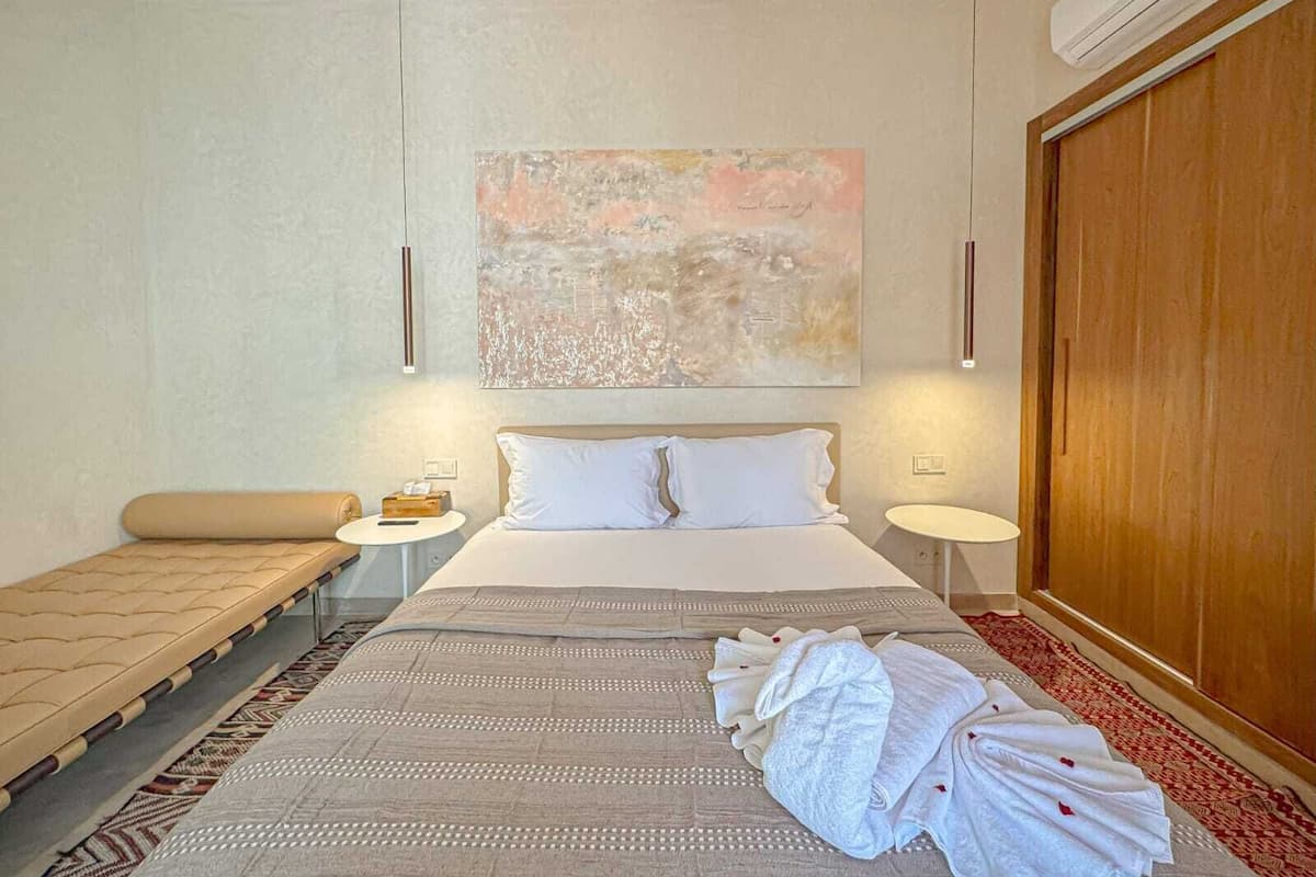 A modern hotel room with a large bed, bedside tables, a bench, and a large abstract painting on the wall.