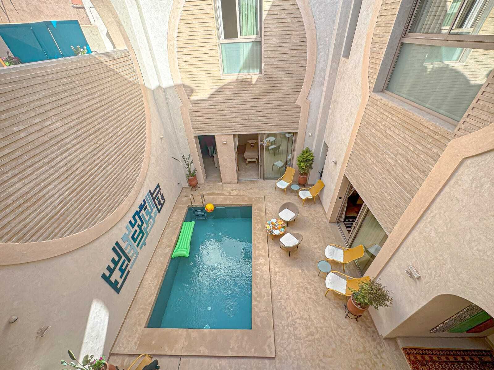An indoor swimming pool with a green inflatable float, surrounded by yellow lounge chairs and potted plants.