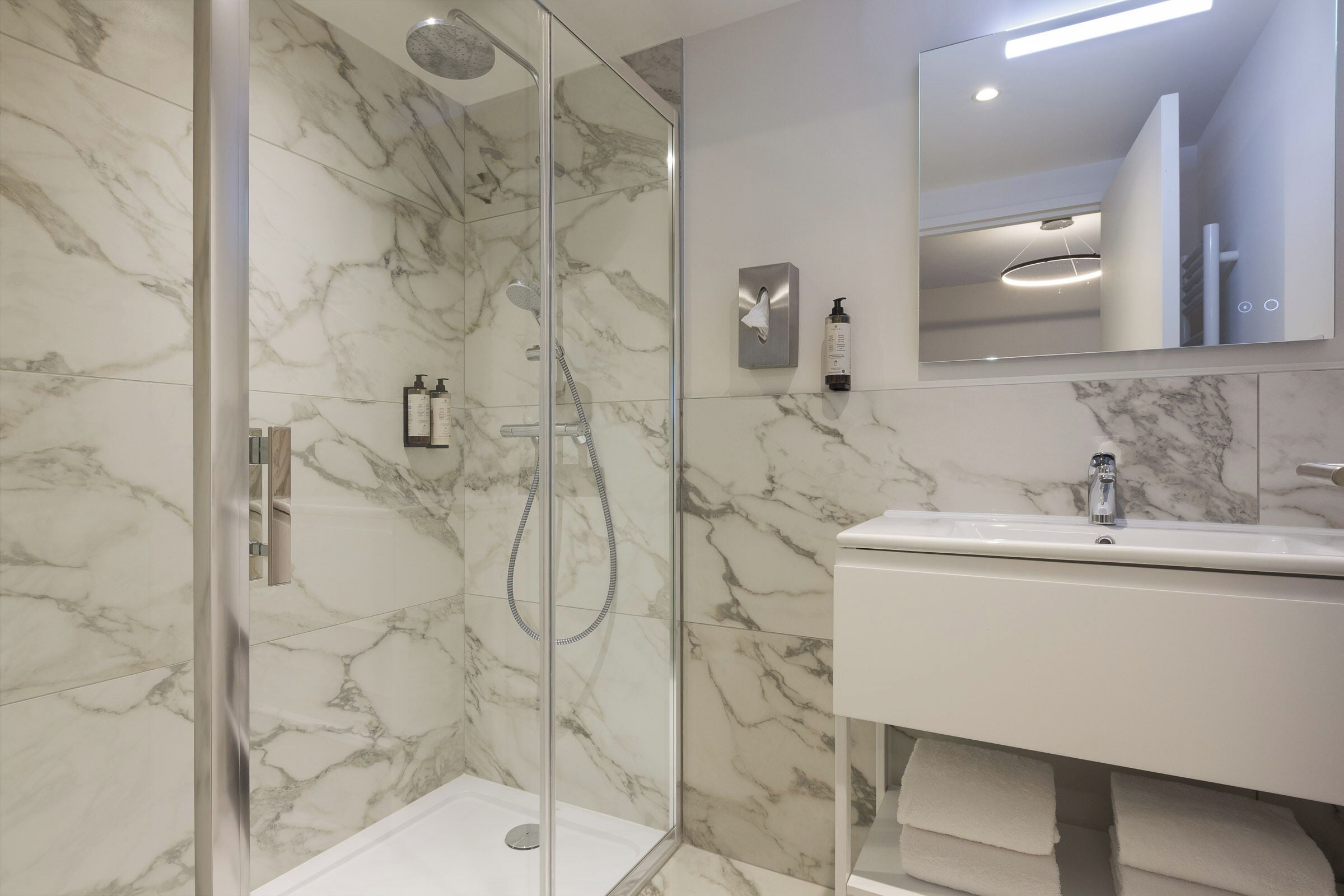 A modern bathroom with a glass-enclosed shower, a marble-tiled wall, a white sink, and a mirror above the sink.