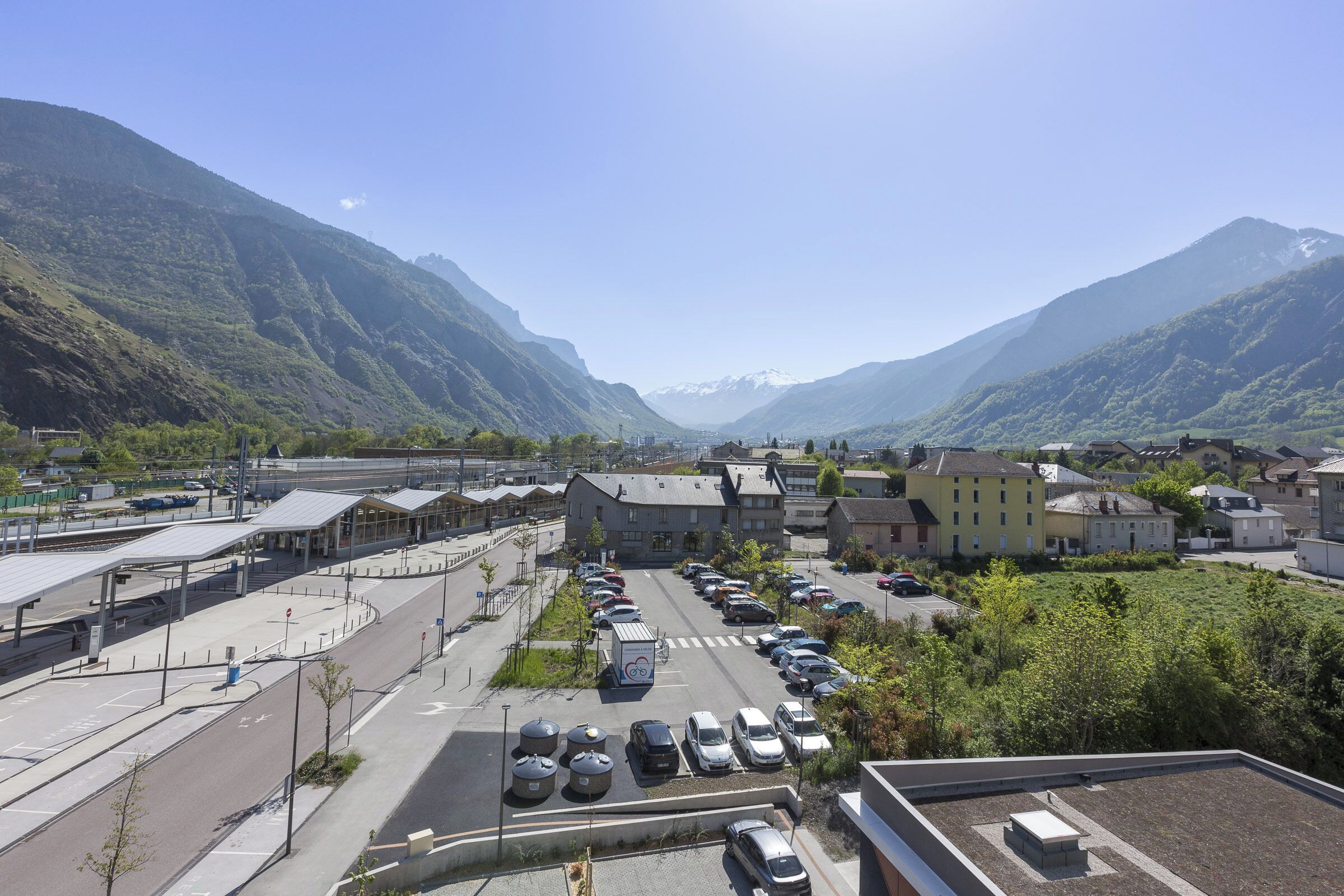 A parking lot with cars, a train station, and buildings in a valley with mountains in the background.