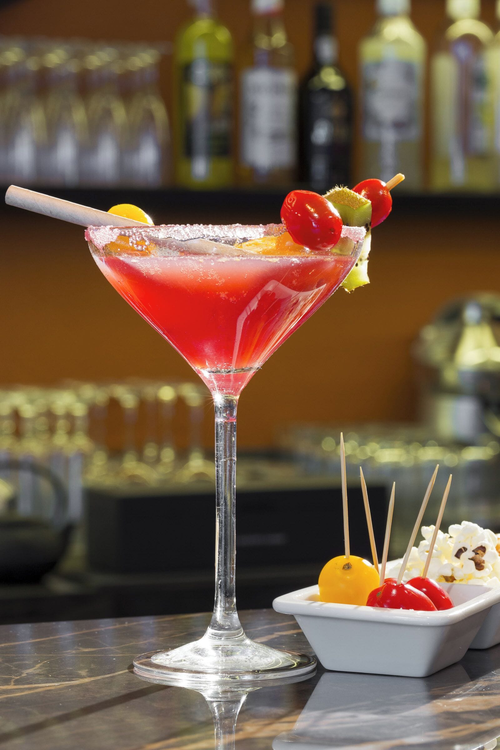 A cocktail in a martini glass with a salted rim, garnished with a cherry, lemon slice, and a skewer with a cherry and a small orange.