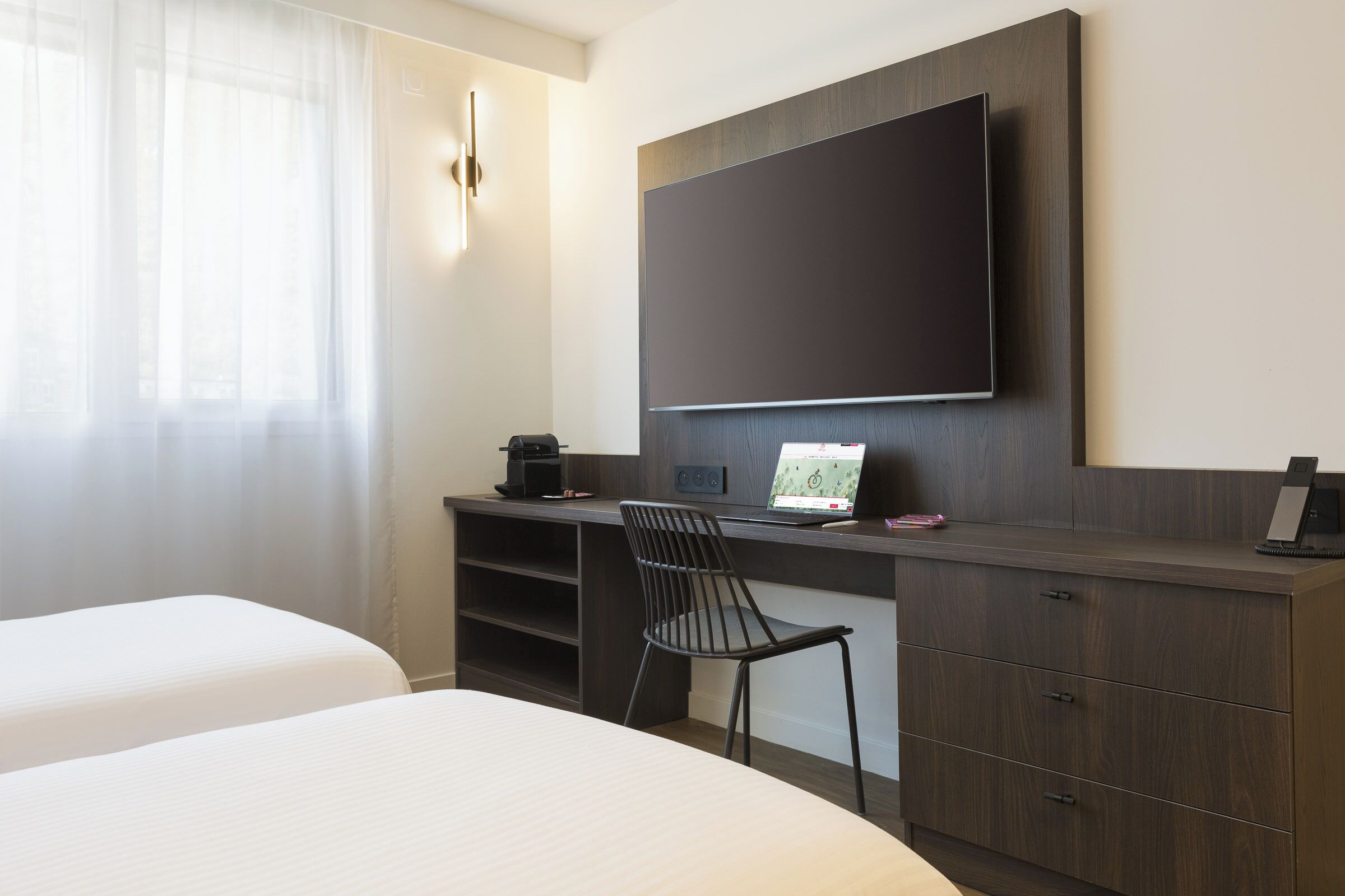 A hotel room with a bed, a desk with a laptop, a chair, a flat-screen TV, and a coffee maker.