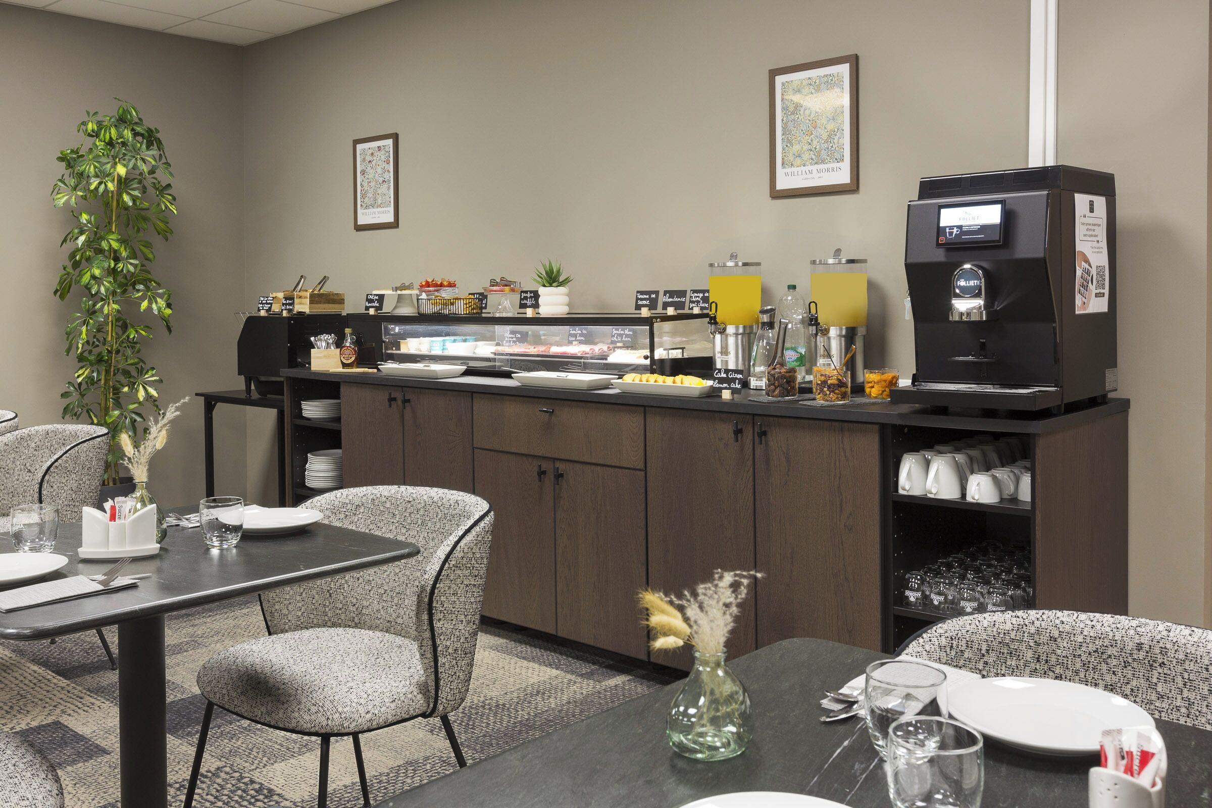 Habitación de hotel con kitchenette, zona de comedor con mesa y sillas, cafetera y planta.