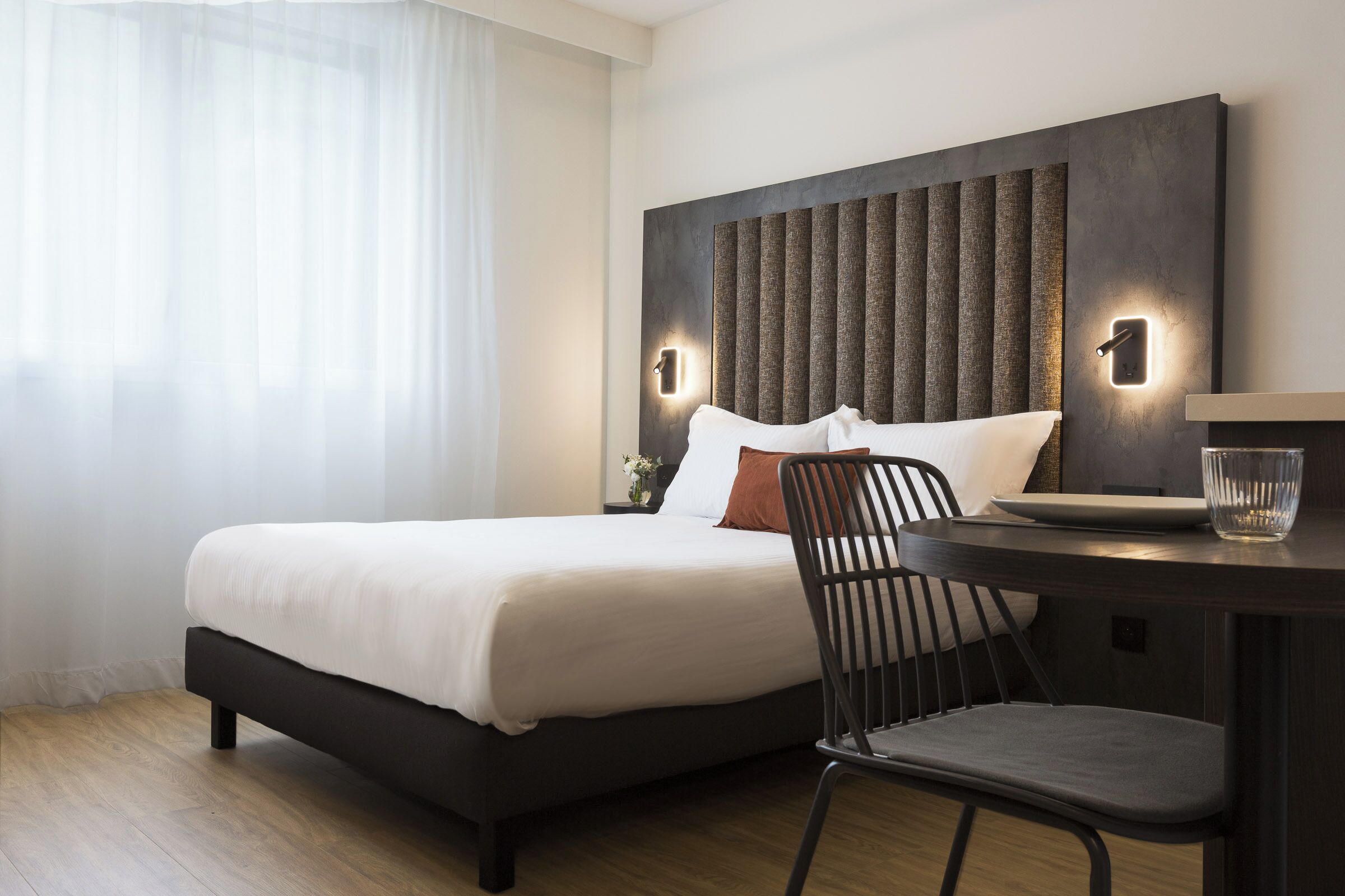 Una habitación de hotel moderna con una cama grande, un escritorio con silla y un ventanal con cortinas translúcidas.