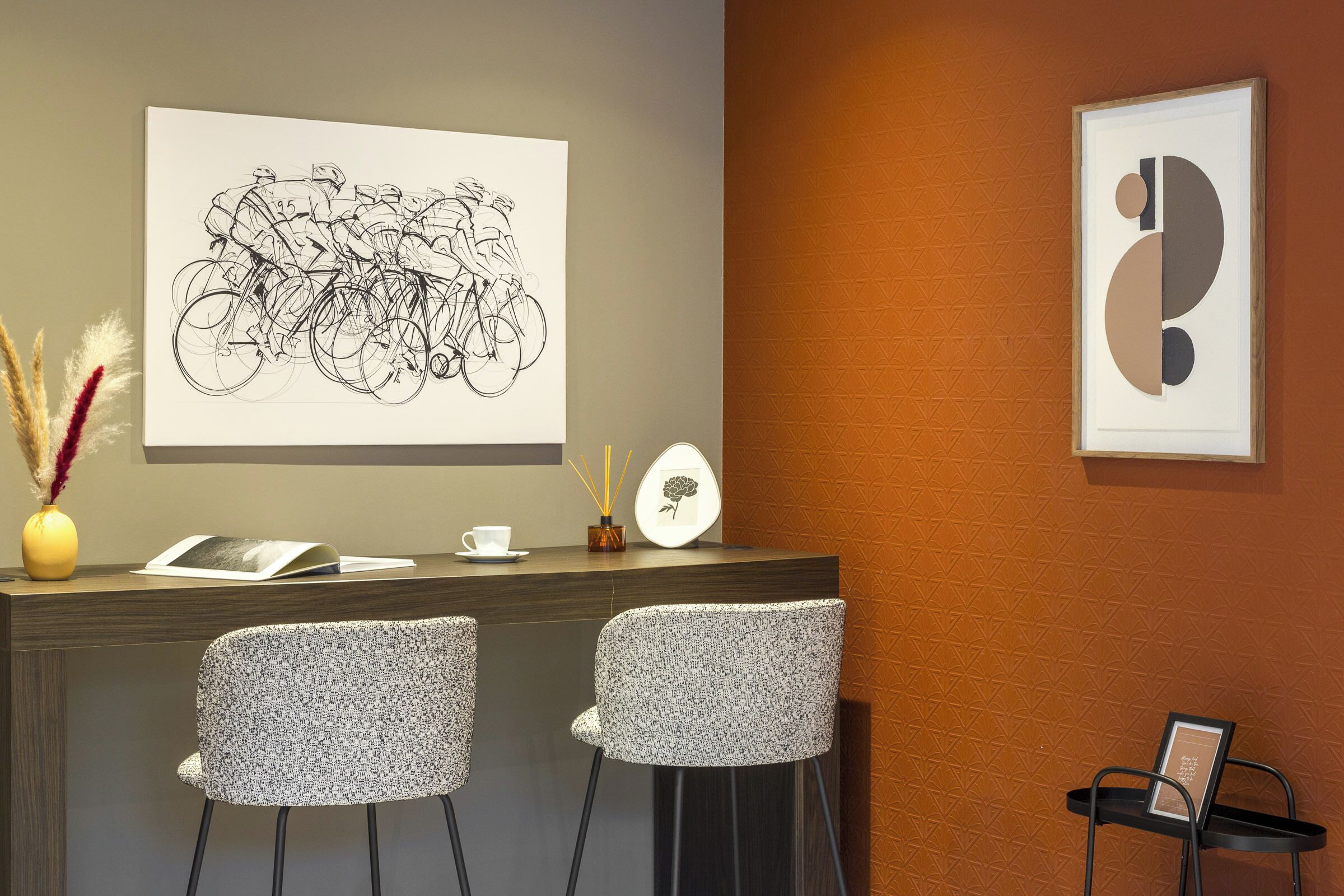 A reception area with a desk, two patterned chairs, a vase with dried plants, a framed abstract artwork, and a wall-mounted picture.