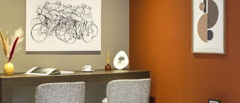 A reception area with a desk, two patterned chairs, a vase with dried plants, a framed abstract artwork, and a wall-mounted picture.