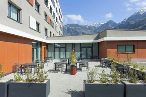 A modern outdoor seating area with tables and chairs, surrounded by planters and overlooking a mountainous landscape. - Nehô Suites St Jean de Maurienne (Saint-Jean-de-Maurienne)