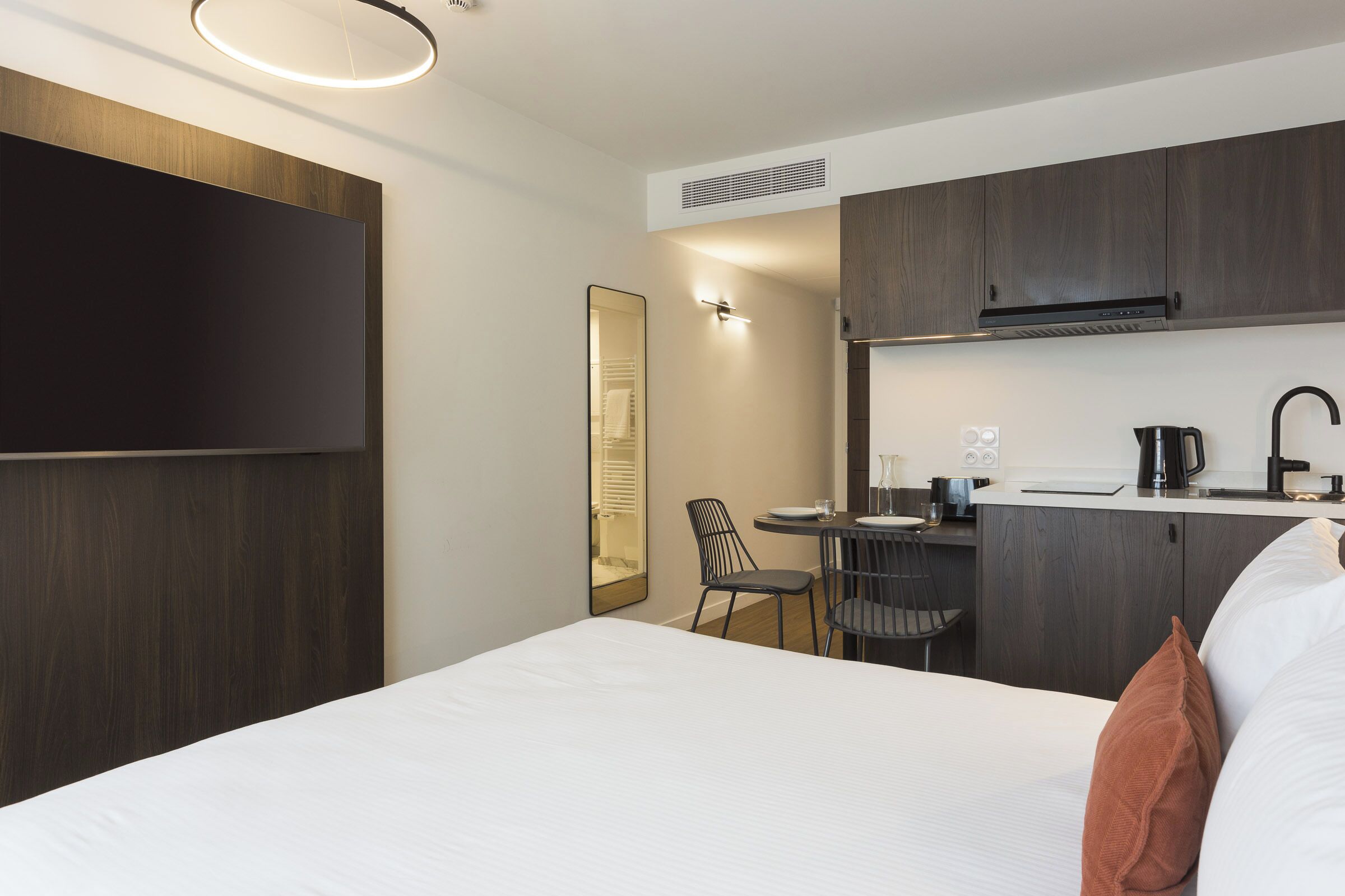 A hotel room with a bed, a desk, a chair, a flat-screen TV, and a kitchenette.