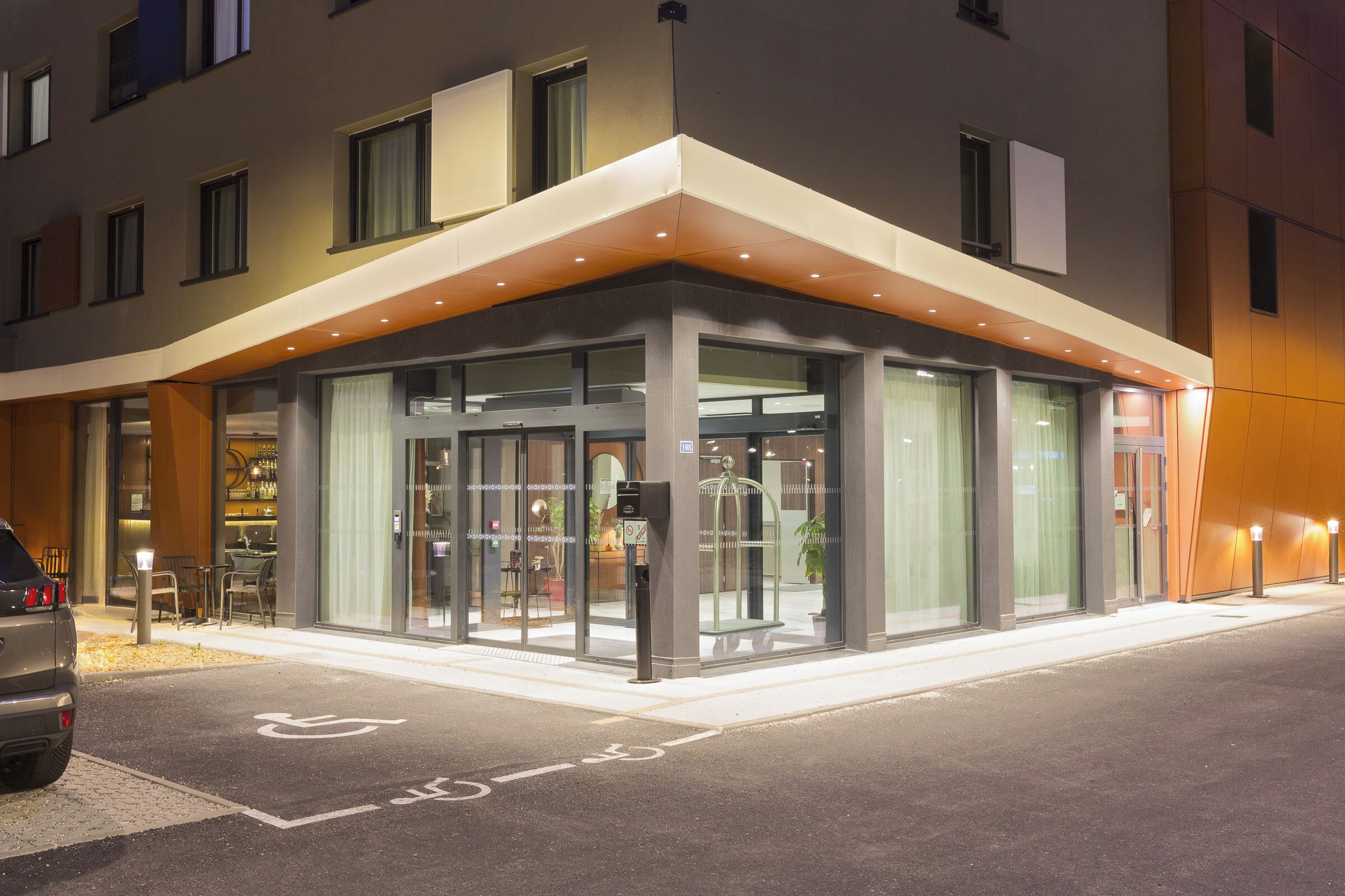 Une entrée d’hôtel moderne avec une porte vitrée, une rampe d’accès pour les personnes à mobilité réduite et un espace de stationnement.