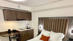 Un dormitorio con cama, mesita de noche, escritorio con silla, zona de cocina con armarios y cabecero empotrado.