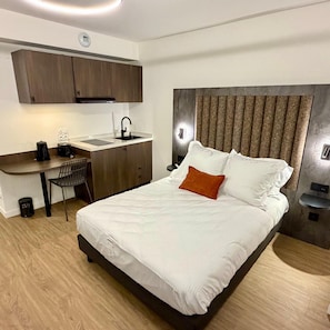 A bedroom with a bed, a nightstand, a bedside table, a desk with a chair, a kitchen area with cabinets, and a built-in headboard. - Nehô Suites St Jean de Maurienne (Saint-Jean-de-Maurienne)