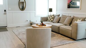 Een moderne woonkamer met een beige bank, een rond pouf en een groot raam dat voor natuurlijk licht zorgt.