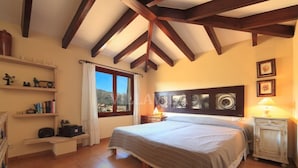 Une chambre avec un grand lit, des poutres en bois, une fenêtre donnant sur un beau paysage, des étagères remplies de livres et une table de chevet avec une lampe.