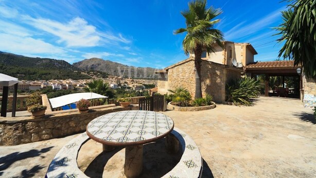 Une terrasse avec une table ronde et des chaises, un palmier et un bâtiment en pierre surmonté d’un toit en tuiles.