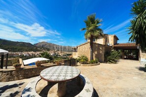 A patio with a round table and chairs, a palm tree, and a stone building with a tiled roof. - Can Coste by Vida Villas Private Pool Mountain Views Walk to Town (Pollença)