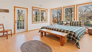 Un dormitorio con una cama grande, un banco y una silla. Varias ventanas ofrecen vistas a árboles y una puerta con un cartel.