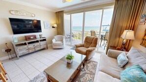 Una sala de estar con televisor de pantalla plana, un sofá, una silla, una mesa de centro y vistas a la playa a través de amplias ventanas.