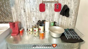 Una cocina con protector de salpicadero de madera, encimera de acero inoxidable y varios utensilios, incluyendo una cafetera roja, un bol blanco y una estantería metálica.