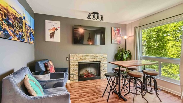A living room with a fireplace, a sofa, a dining table with stools, and a large window with a view of trees. - Music City Living - 2BR/2BA Riverfront Paradise with FREE PARKING (Nashville)