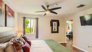 A bedroom with a bed, a ceiling fan, a window with curtains, a TV, and a view into another room. - Music City Living - 2BR/2BA Riverfront Paradise with FREE PARKING (Nashville)