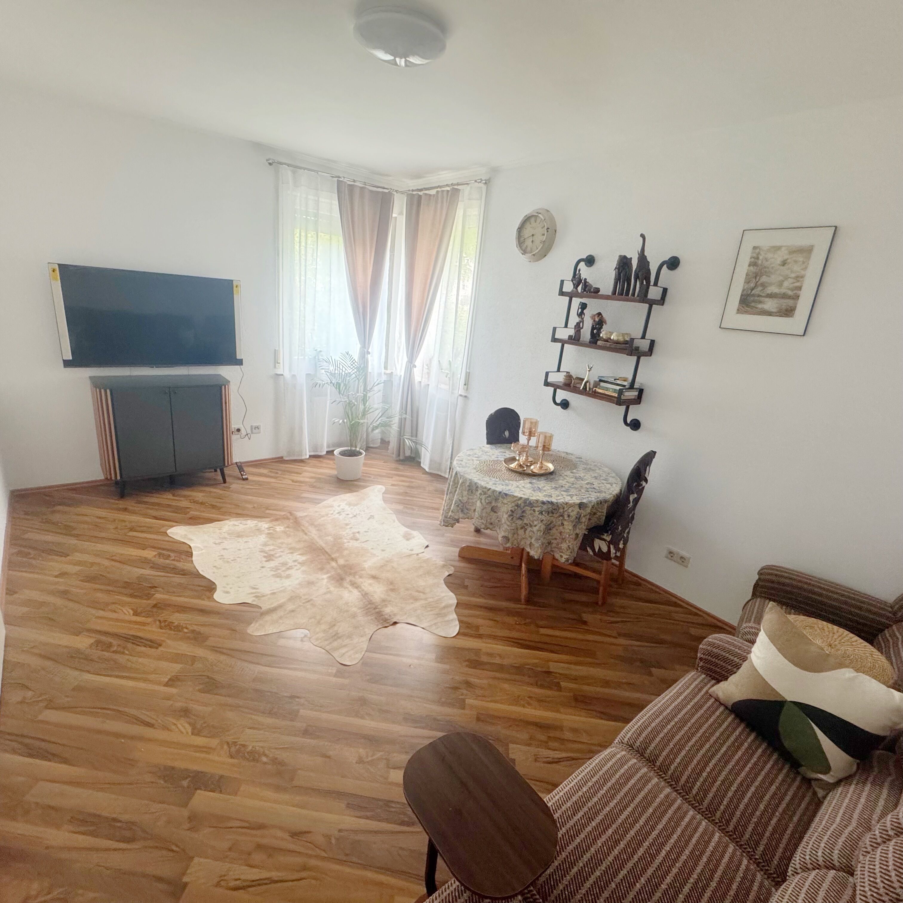 Een woonkamer met houten vloer, een bank, een televisie, een eettafel met stoelen en een raam met gordijnen.