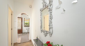 Un pasillo con piso de madera, pared blanca, un espejo decorativo y una planta en maceta con flores rojas.