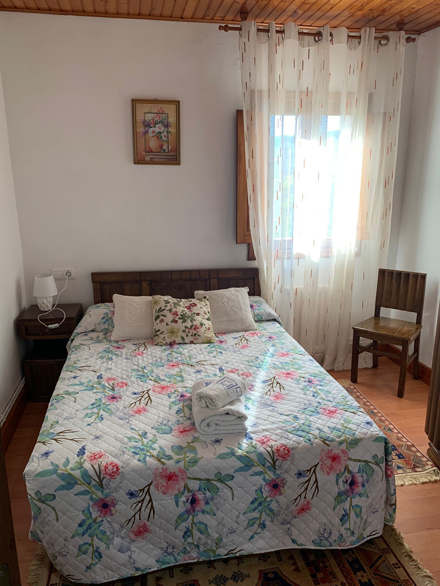A bedroom with a floral bedspread, a wooden headboard, a nightstand with a lamp, a chair, and a window with sheer curtains.