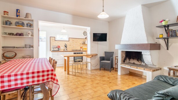 Un salon avec une cheminée, un coin repas avec une nappe à carreaux et une cuisine visible à travers une porte ouverte.