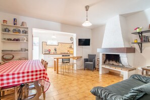 Een woonkamer met een open haard, een eethoek met geruit tafelkleed, en een keuken die door een open deur zichtbaar is.