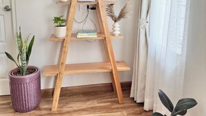 A wooden ladder shelf with books and decorative items, flanked by potted plants, in a room with wooden flooring and a white door. - Peaceful 2BHK Retreat | Near Forest Reserve (Kathmandu)
