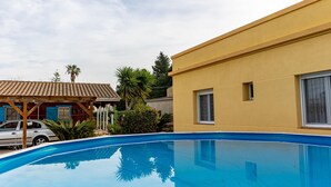 En swimmingpool med fliseterrasse, en gul bygning med et vindue, og en rød bygning med flisebelagt tag i baggrunden.